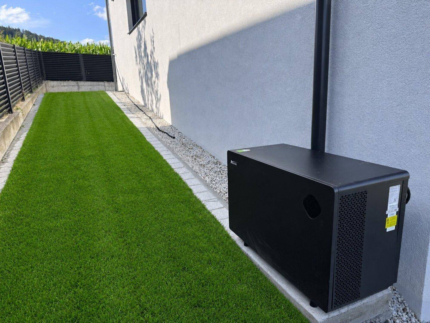 Schwarze Wärmepumpe oder Klimaanlage an der Hauswand neben einem schmalen Rasenstreifen.