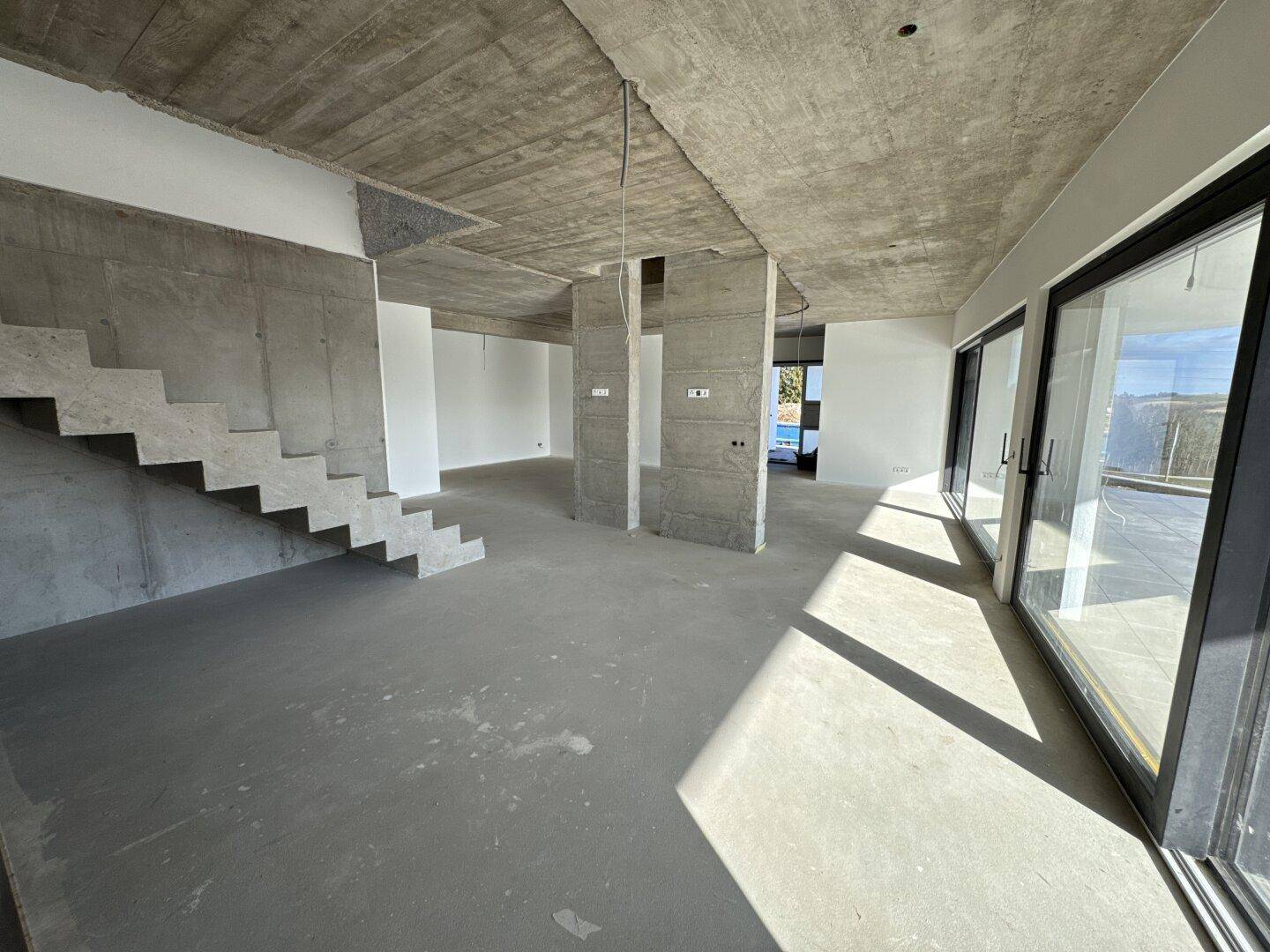 Großer offener Raum im Rohbau mit Betontreppe, hohen Decken und viel Tageslicht.
