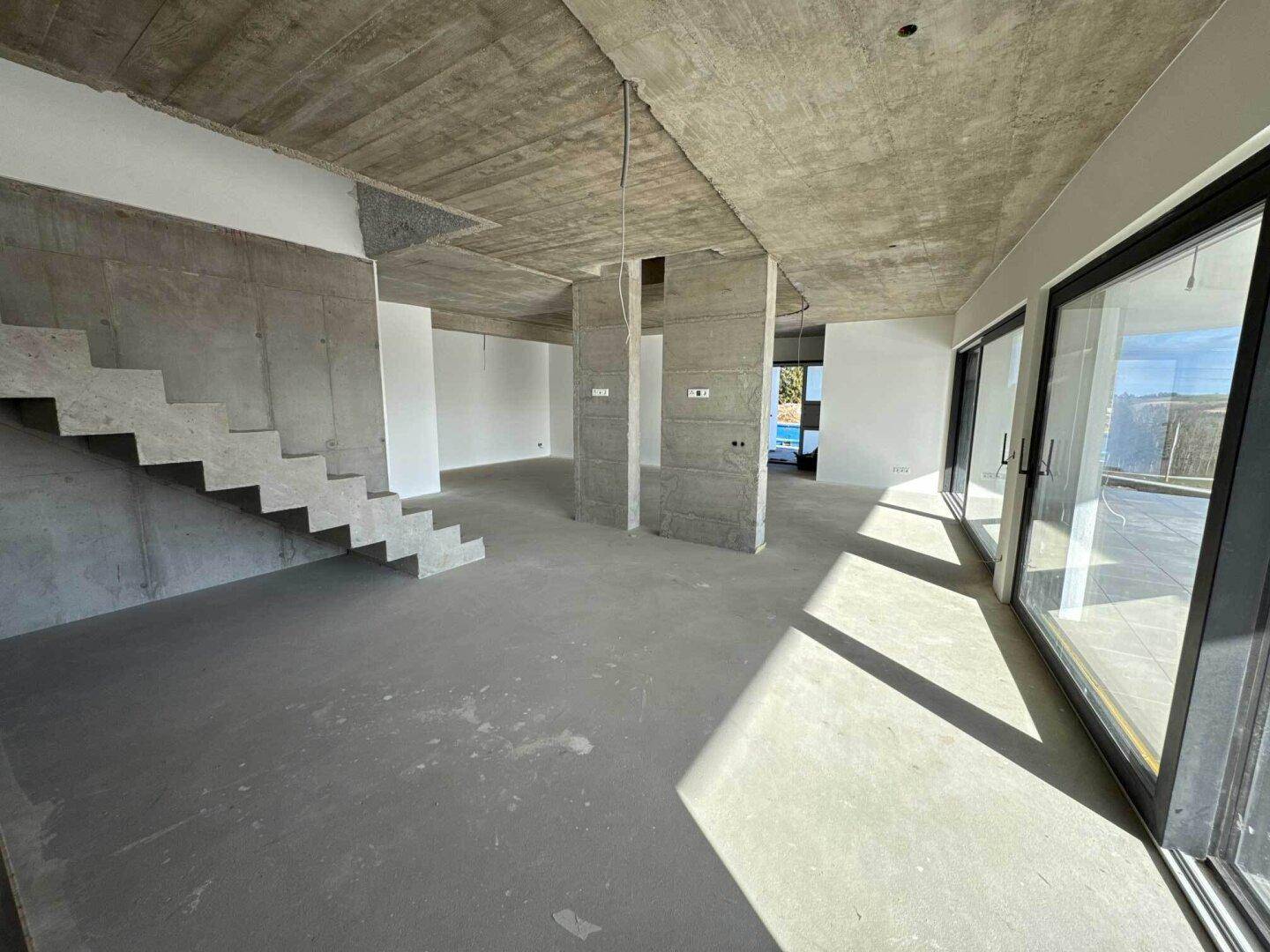 Offener Wohnbereich im Rohbau mit Betontreppe, Sichtbetonwänden und großen Fensterfronten.