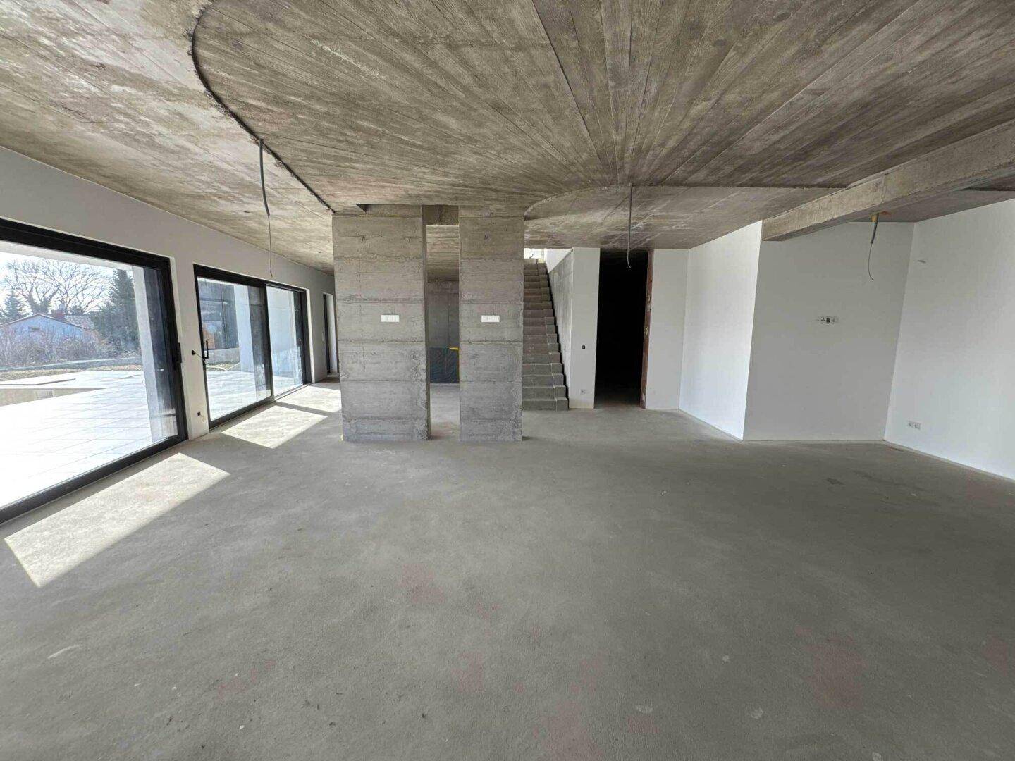 Großer offener Wohnbereich im Rohbau mit Betonsäulen und Zugang zur Außenterrasse.