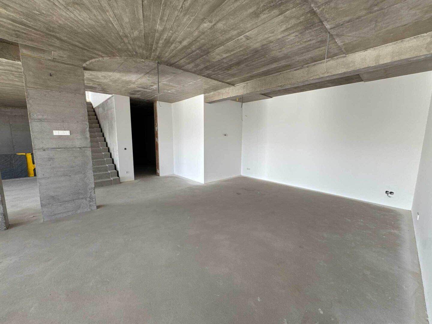 Geräumiger offener Raum im Rohbau mit Sichtbetondecke und weißen Wänden, bereit für den Ausbau.