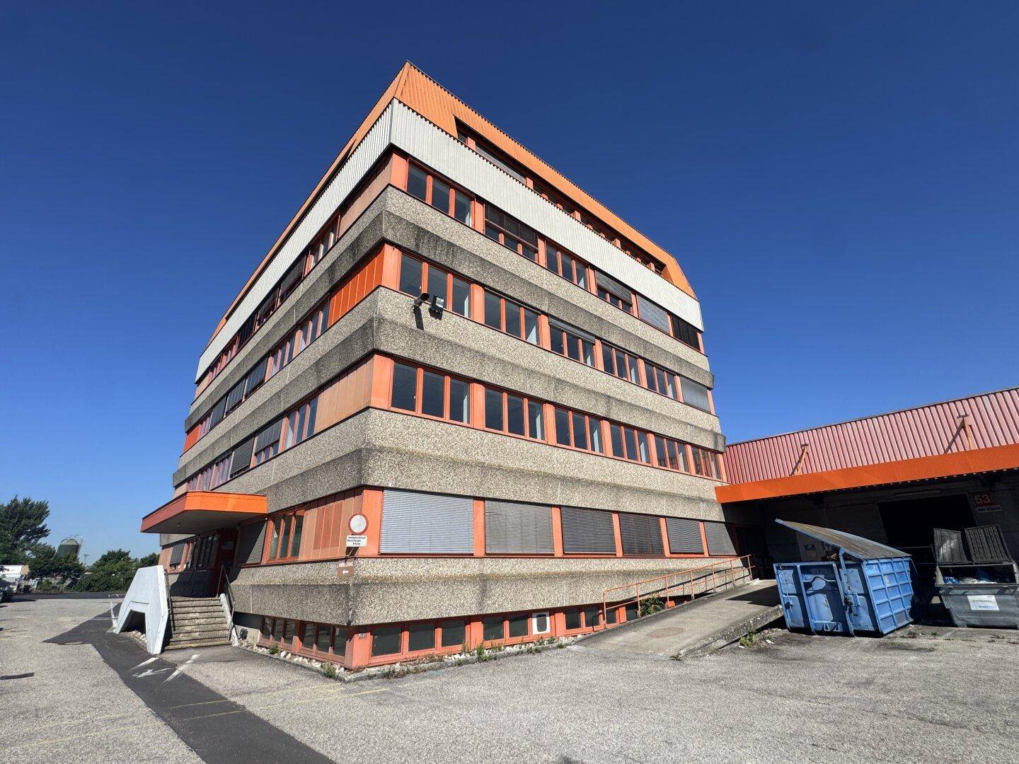 Gesamtansicht des Bürogebäudes mit Parkplatz und Laderampenbereich unter strahlend blauem Himmel.