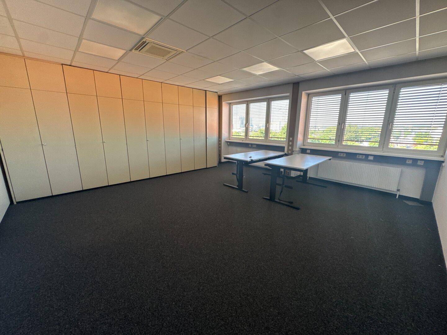 Büro mit Einbauschränken entlang einer Wand, dunklem Teppichboden und Fenstern mit Jalousien.