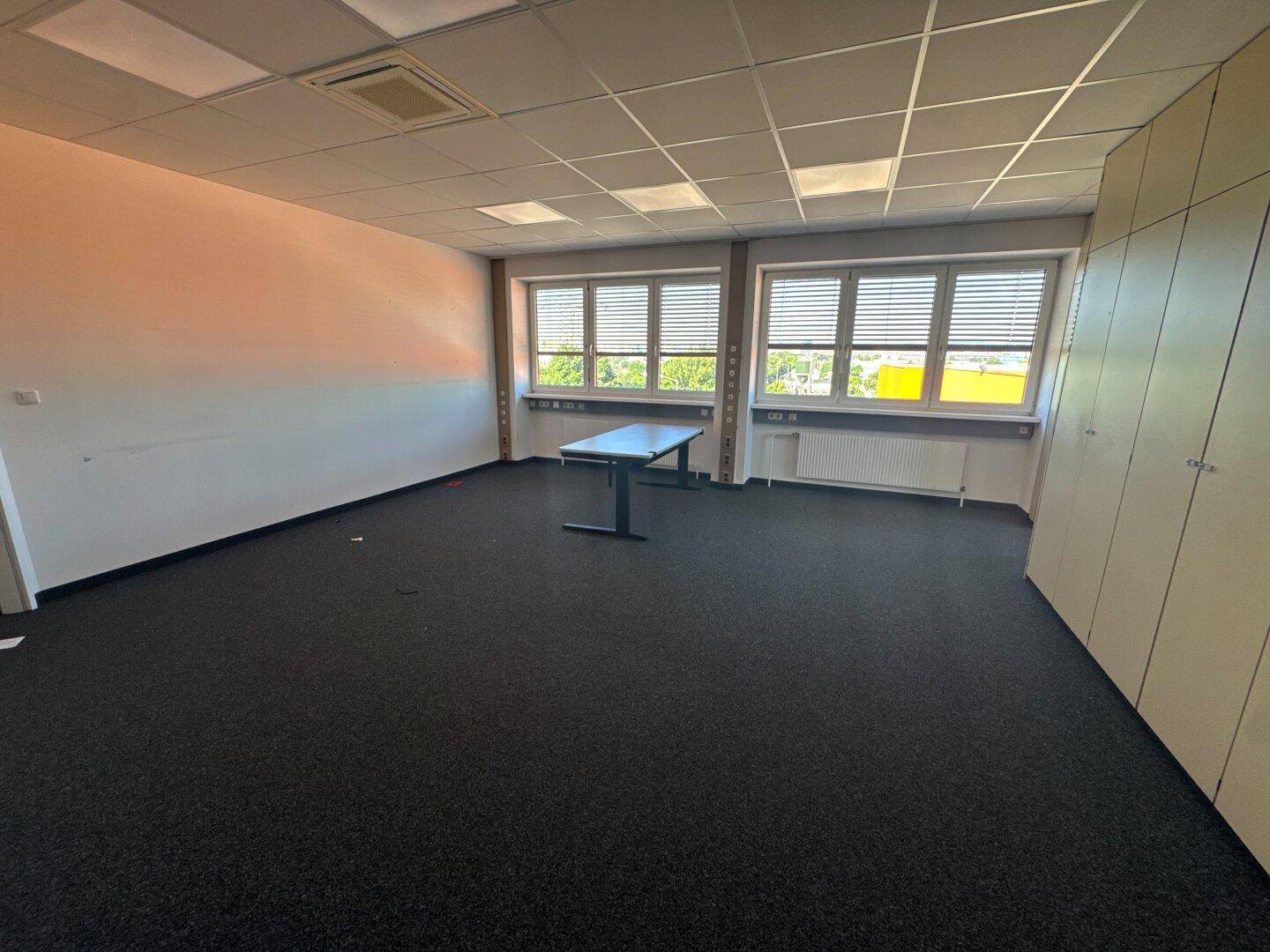 Geräumiges Büro mit einem Tisch, dunklem Teppichboden und Fenstern mit Jalousien.