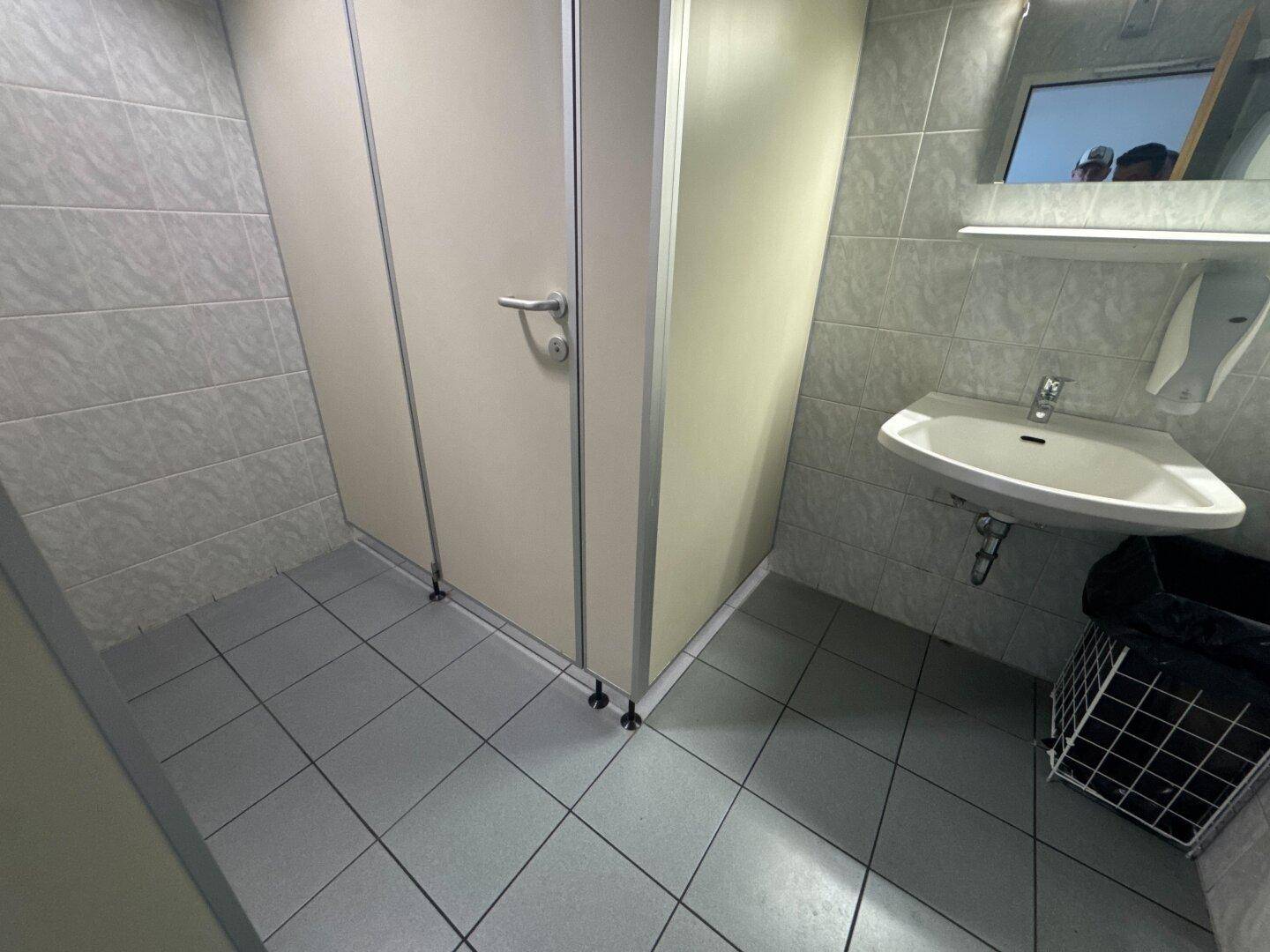Einfache Toilettenkabinen mit Fliesenboden und Waschbecken in einem öffentlichen Sanitärbereich.