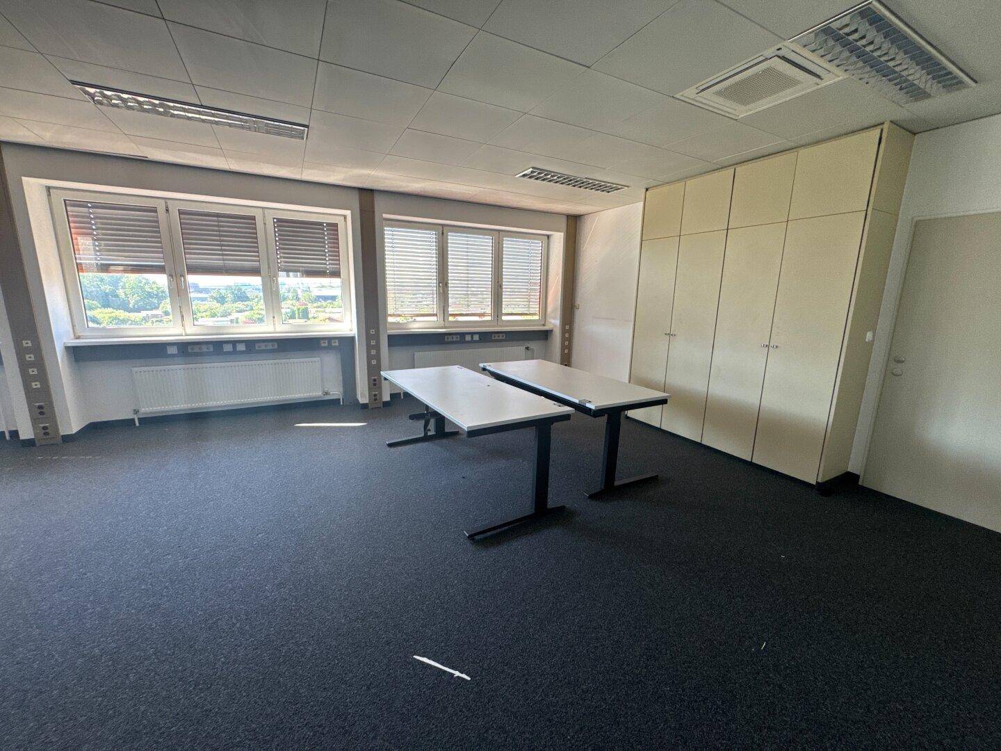 Büro mit Einbauschränken, zwei Tischen und Fenstern mit Jalousien, gut beleuchtet.