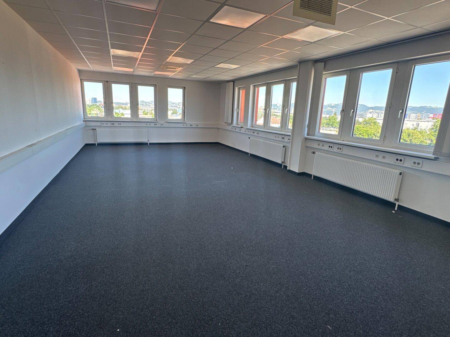 Großes, leeres Büro mit dunklem Teppichboden und vielen Fenstern, die eine weite Aussicht bieten.