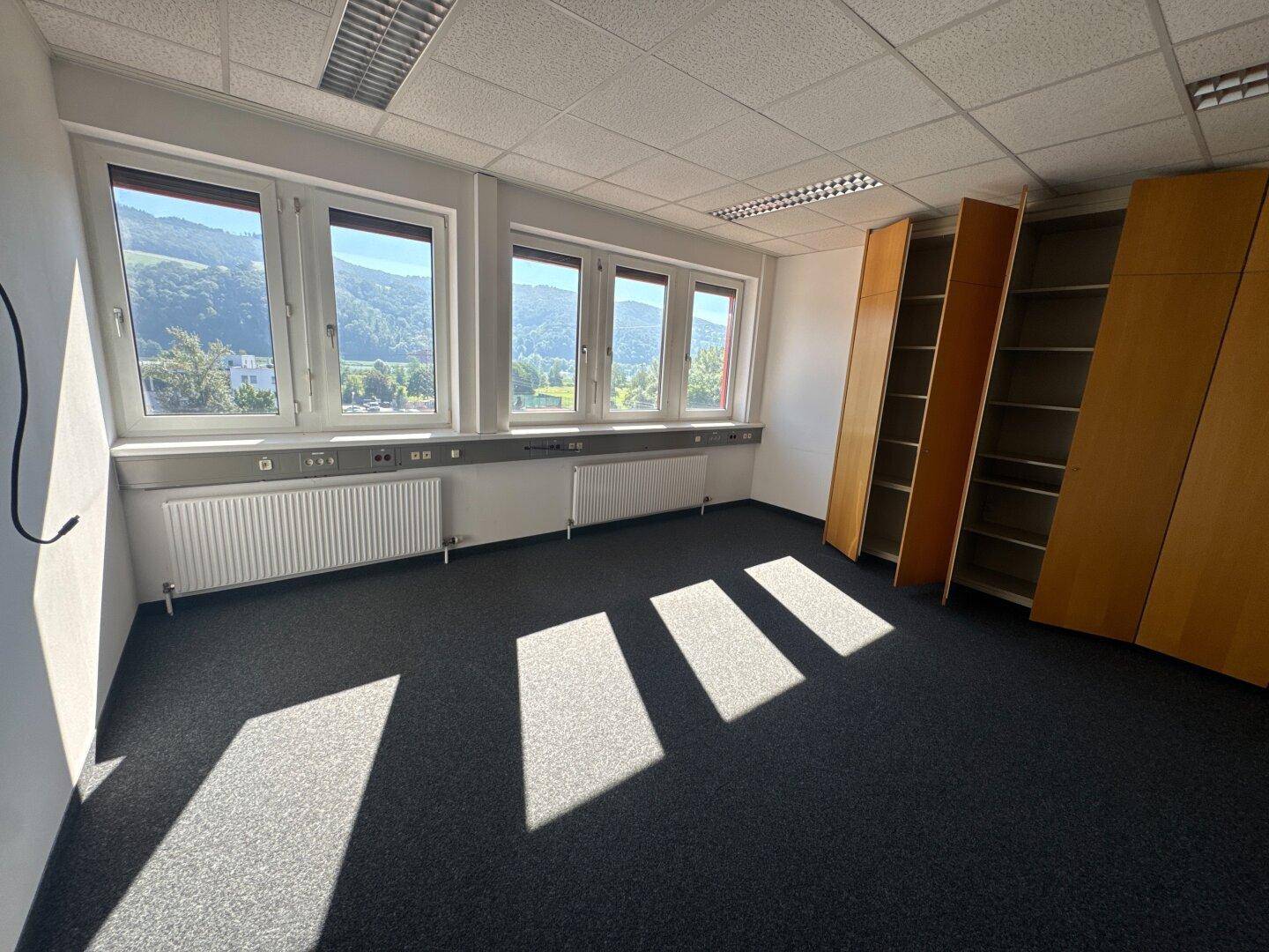 Büro mit dunklem Teppichboden, leeren Regalen und Fenstern mit weitem Ausblick.
