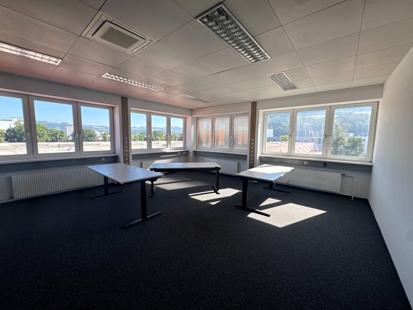 Helles Büro mit mehreren Tischen, dunklem Teppichboden und weitem Ausblick durch die Fenster.