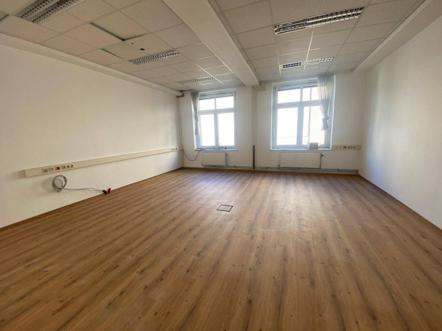 Leeres Büro mit Laminatboden, großen Fenstern und weißen Wänden, bereit zur individuellen Gestaltung.