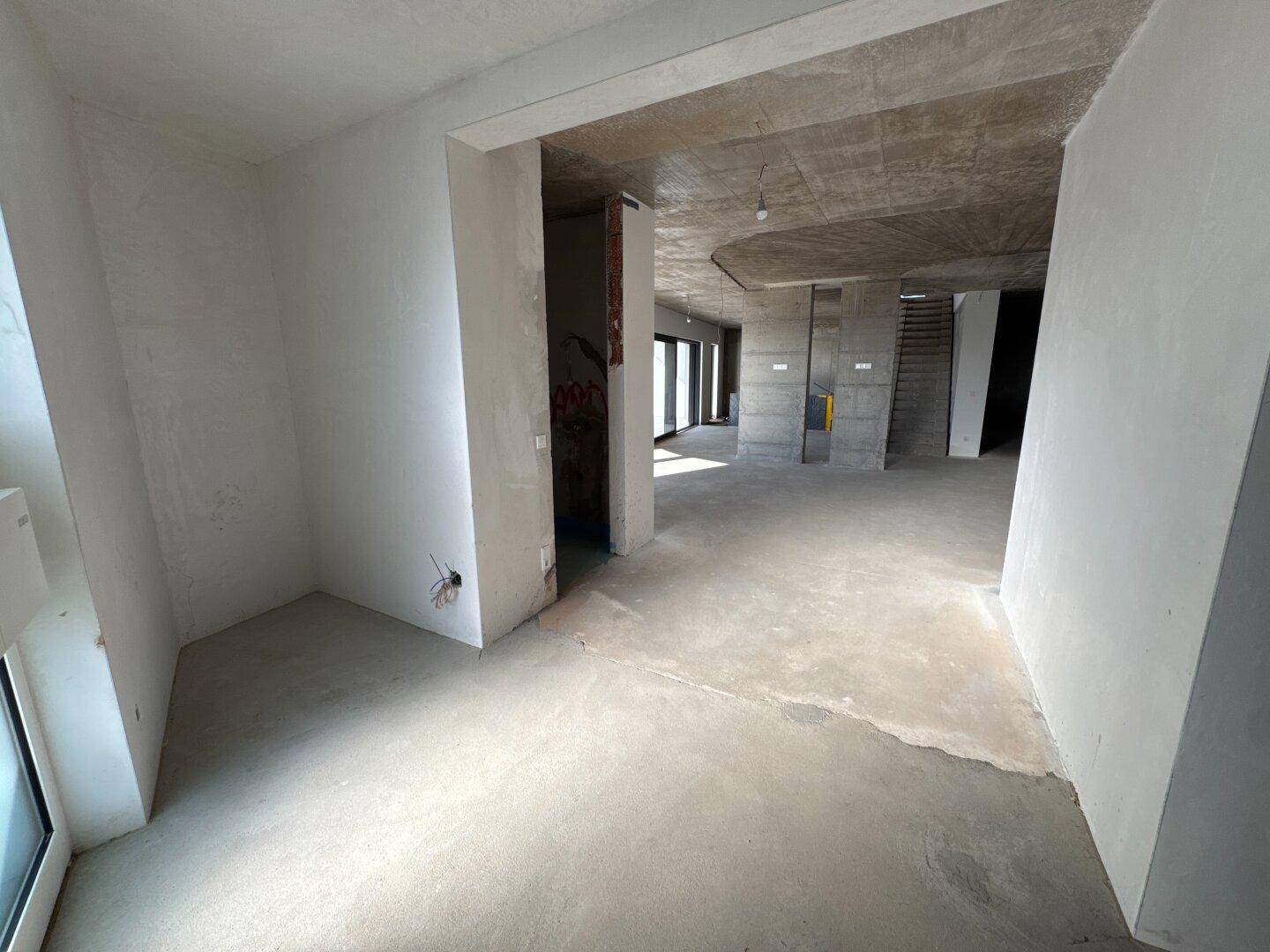Geräumiger Wohnbereich im Rohbau mit Betonboden und großen Fensterfronten zum Garten.
