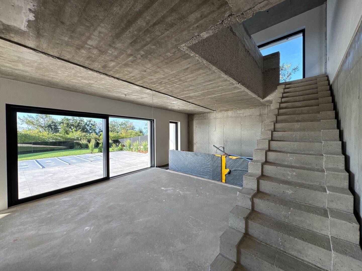 Offener Wohnbereich im Rohbau mit Betontreppe und großen Schiebetüren zur Terrasse.