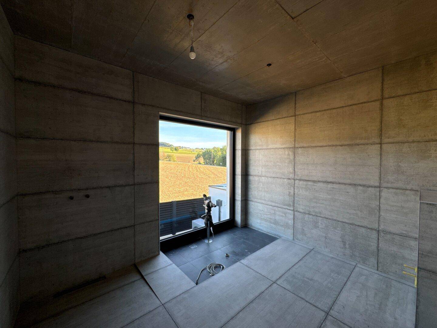 Schlafzimmer im Rohbau mit Sichtbetonwänden, Fliesenboden und großem Fenster mit Landschaftsblick.
