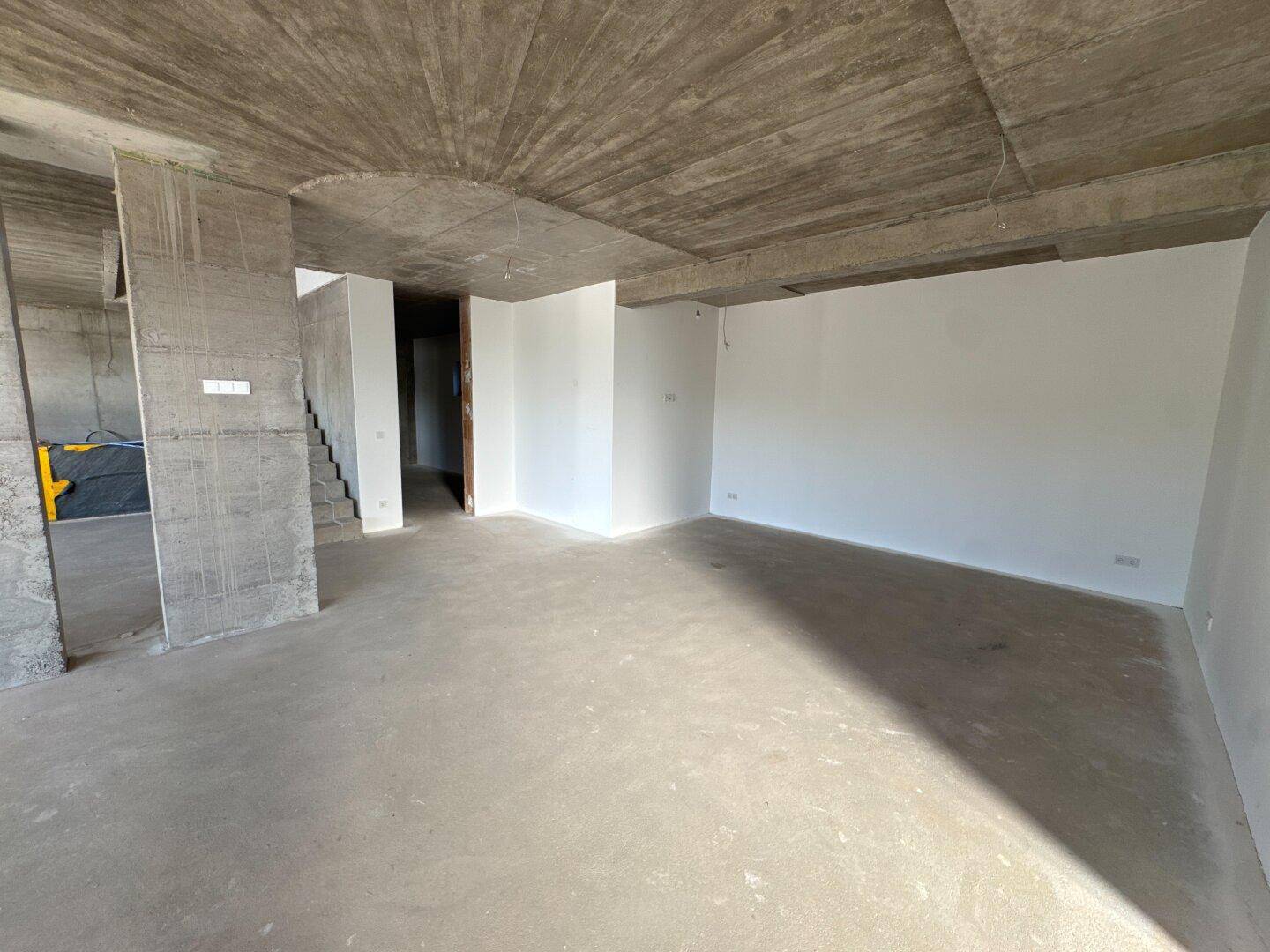 Geräumiger Wohnbereich im Rohbau mit unverputzter Betondecke und teilweise verputzten Wänden.