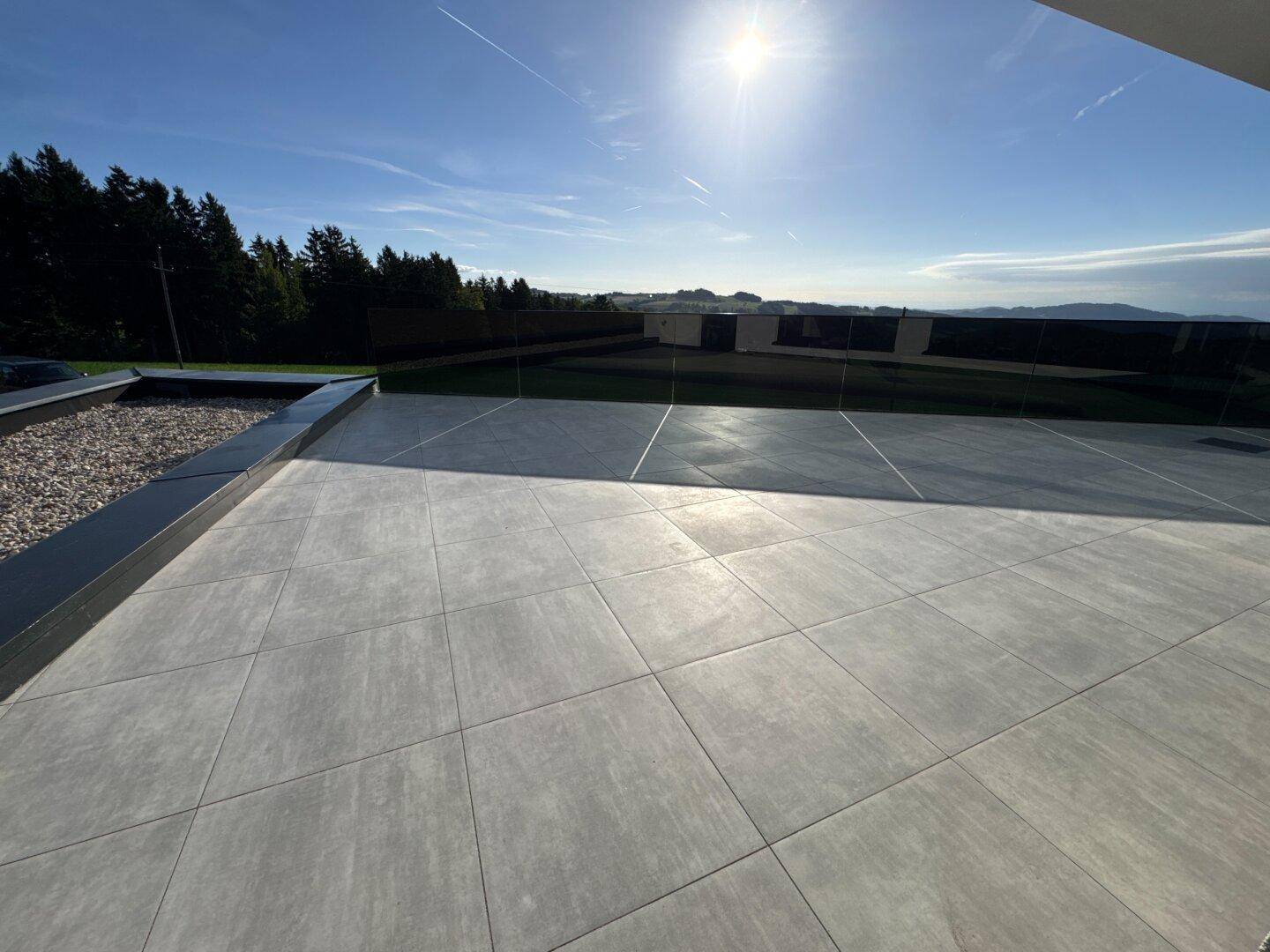 Sonnige Terrasse mit großformatigen Fliesen und Blick auf die umliegende Natur und den Rasen.