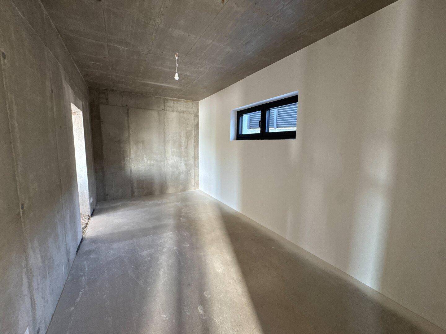 Schlafzimmer im Rohbau mit Betonwänden, einer verputzten Wand und kleinem Fenster.