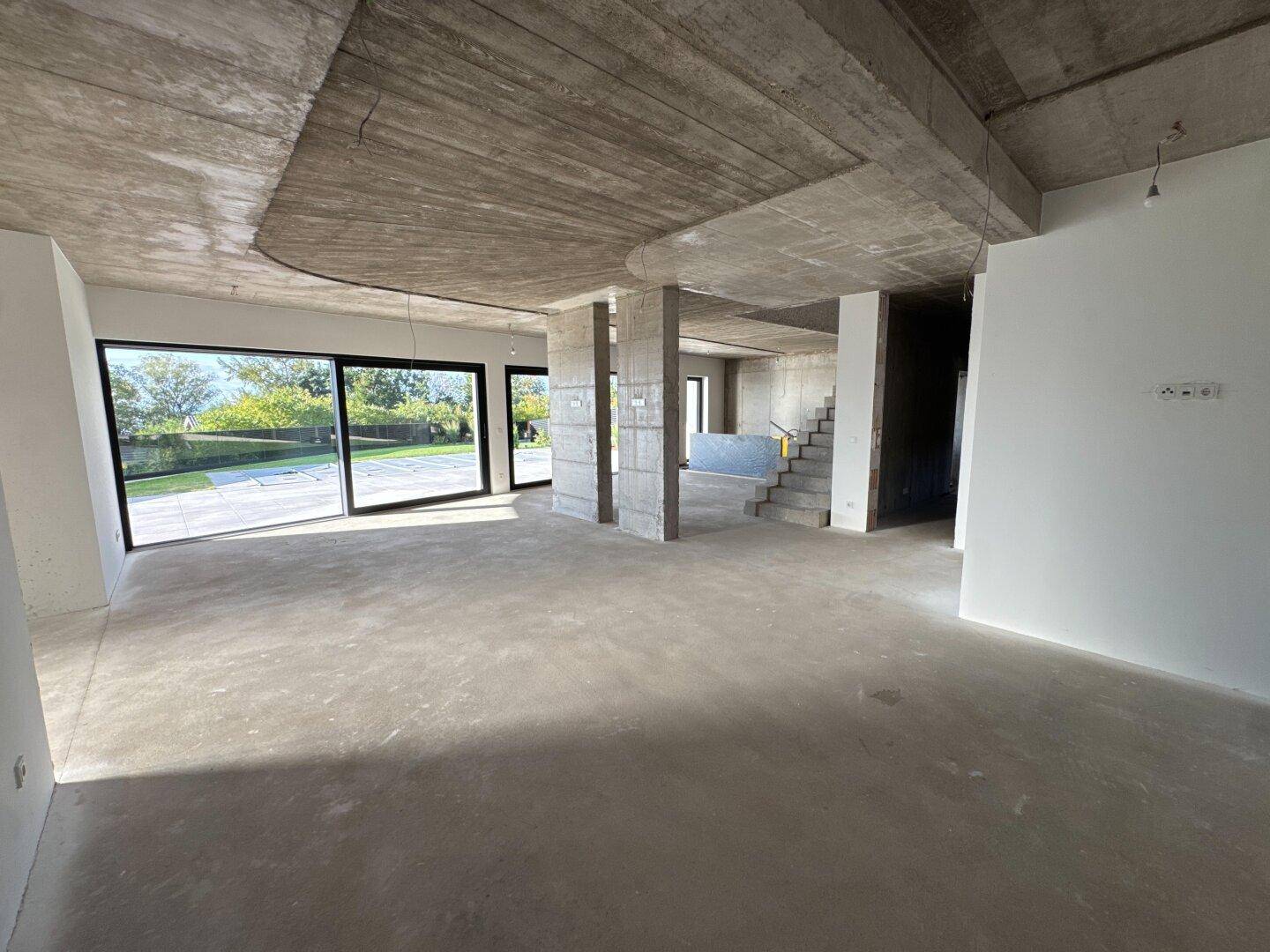 Offener Wohnbereich im Rohbau mit Betondecke, Säulen und Blick auf die Treppe.