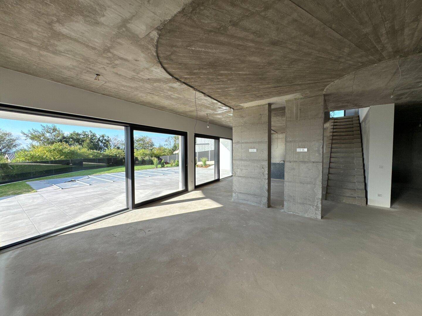 Heller Wohnbereich im Rohbau mit Betondecke und großen Fensterfronten zum Außenbereich.
