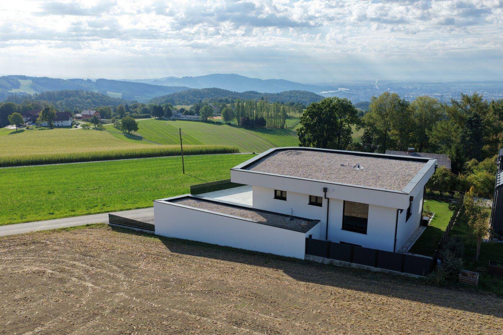Moderne Architektur mit Flachdach und Holzterrasse, umgeben von grünen Feldern und Hügeln.