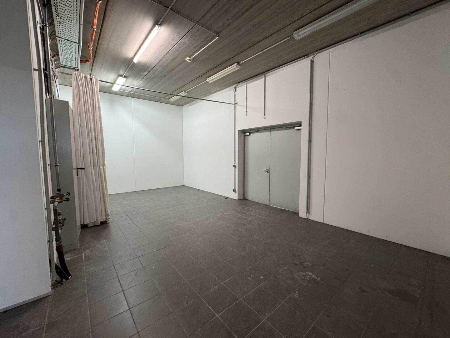 Einfacher Lagerraum mit Fliesenboden, weißen Wänden und sichtbaren Installationen an der Decke.