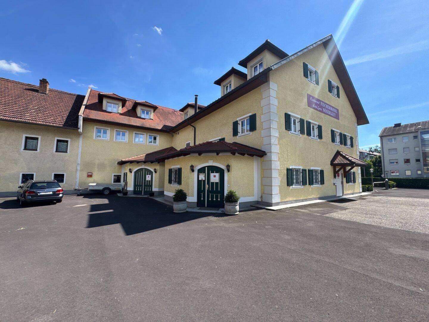 Traditionelles Gebäude mit gelber Fassade, grünen Fensterläden und gepflastertem Vorplatz.
