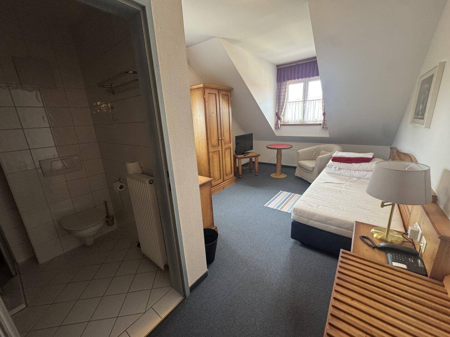 Gemütliches Dachzimmer mit Einzelbett, Holzschrank und angrenzendem Badezimmer.