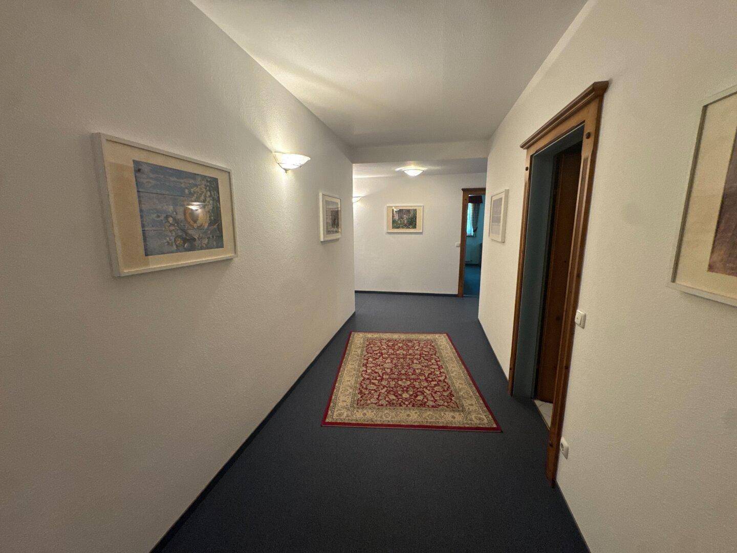 Langer Flur mit Teppichboden, weißen Wänden und Bildern an den Seiten.