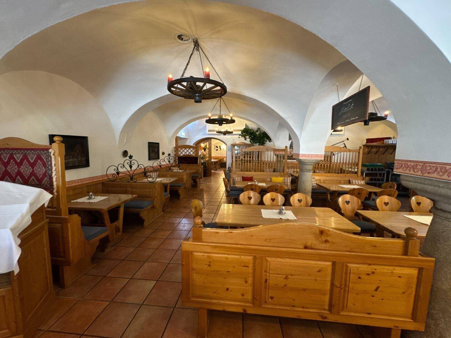 Traditioneller Speisesaal mit Holzbänken, Tischen und gewölbter Decke, beleuchtet von Radleuchtern.