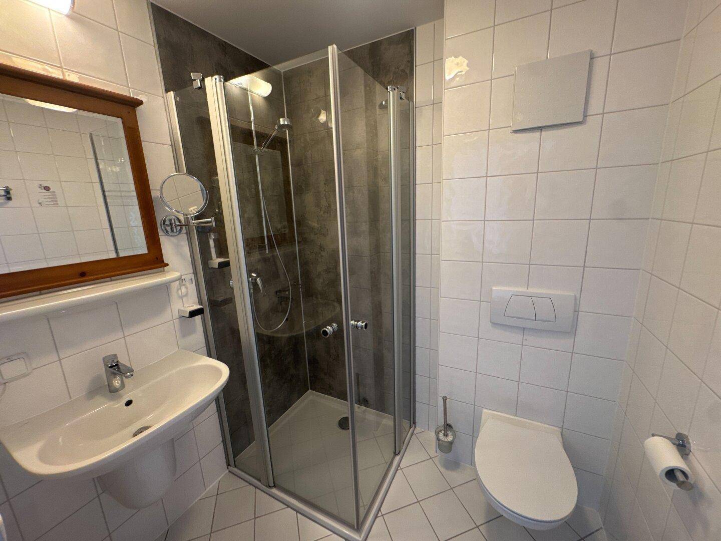 Helles Badezimmer mit Eckdusche, WC, Waschbecken und weißen Fliesen an Wänden und Boden.