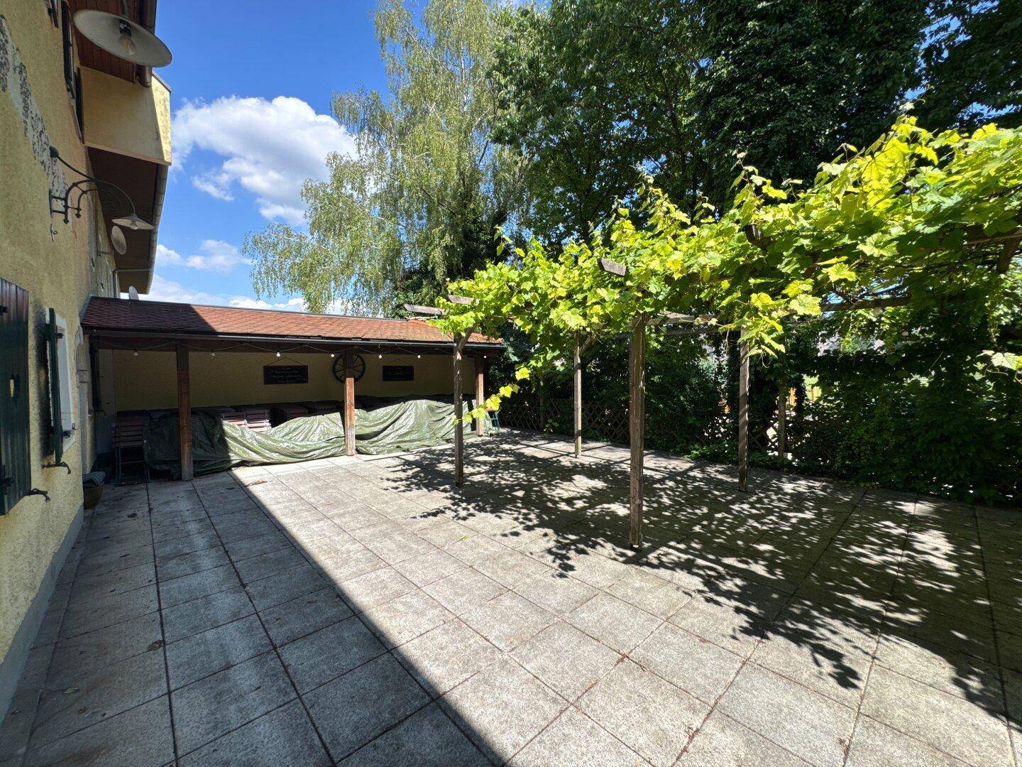 Gepflasterte Terrasse mit überdachtem Bereich und üppigem Grün, ideal für entspannte Stunden im Freien.