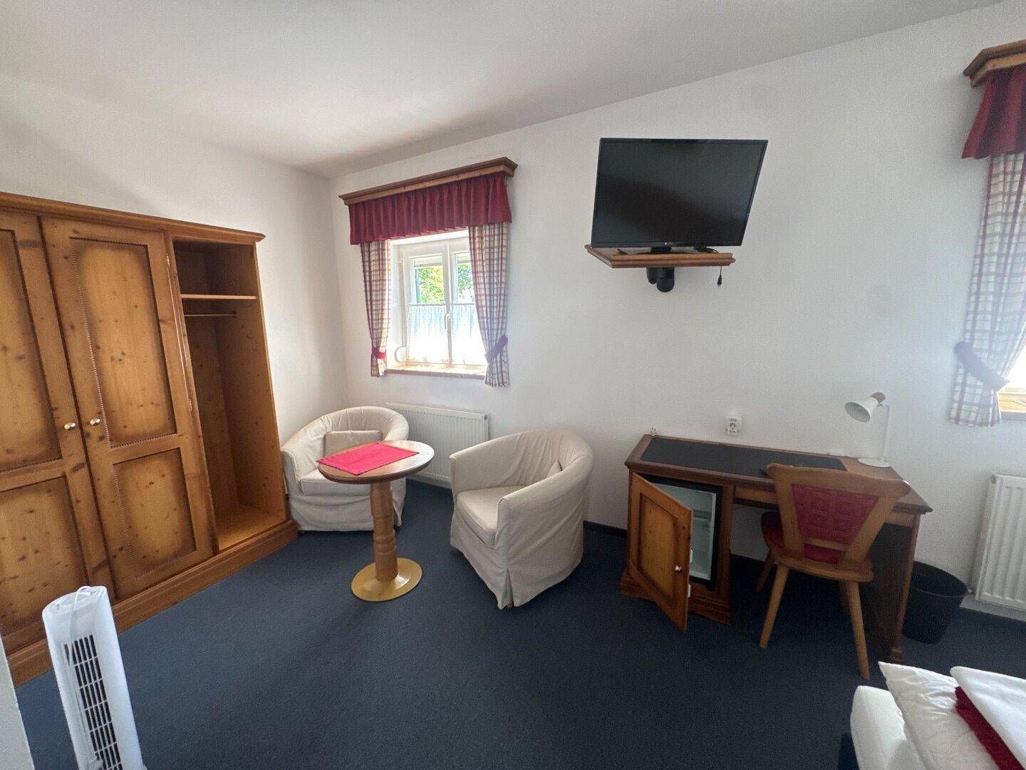 Schlafzimmer mit Holzschrank, zwei Sesseln, Schreibtisch und einem an der Wand montierten Fernseher.