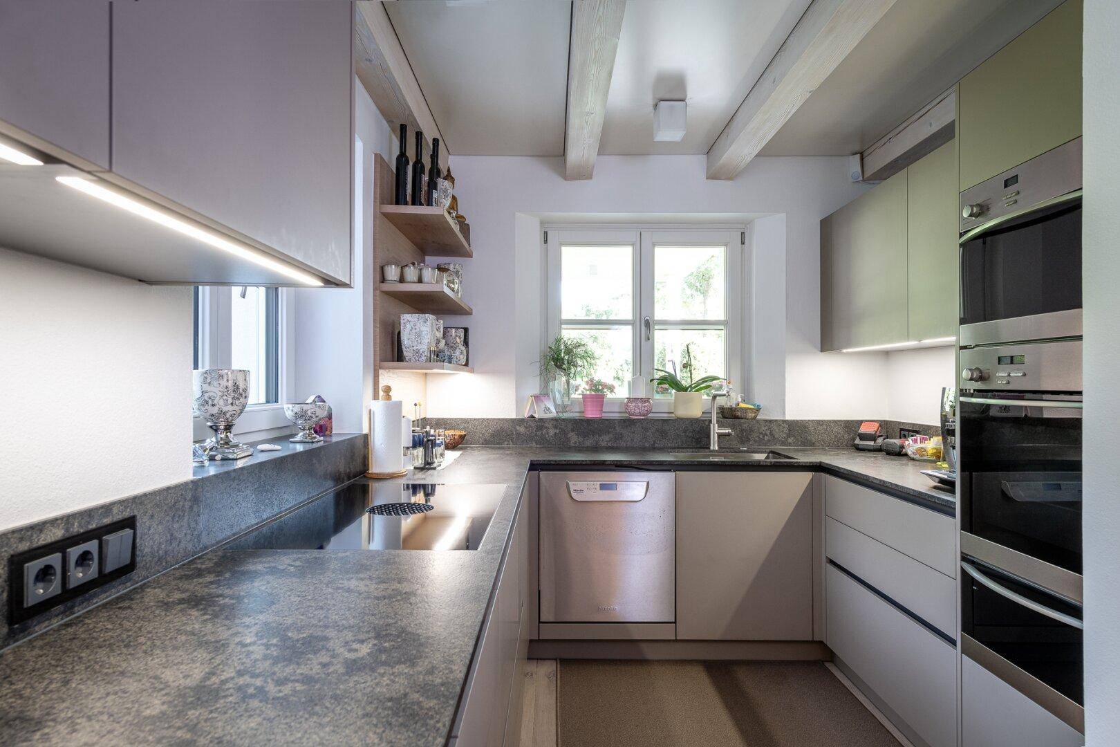 Moderne Küche mit Einbaugeräten, dunkler Arbeitsplatte und Holzbalkendecke, hell durch Fenster.