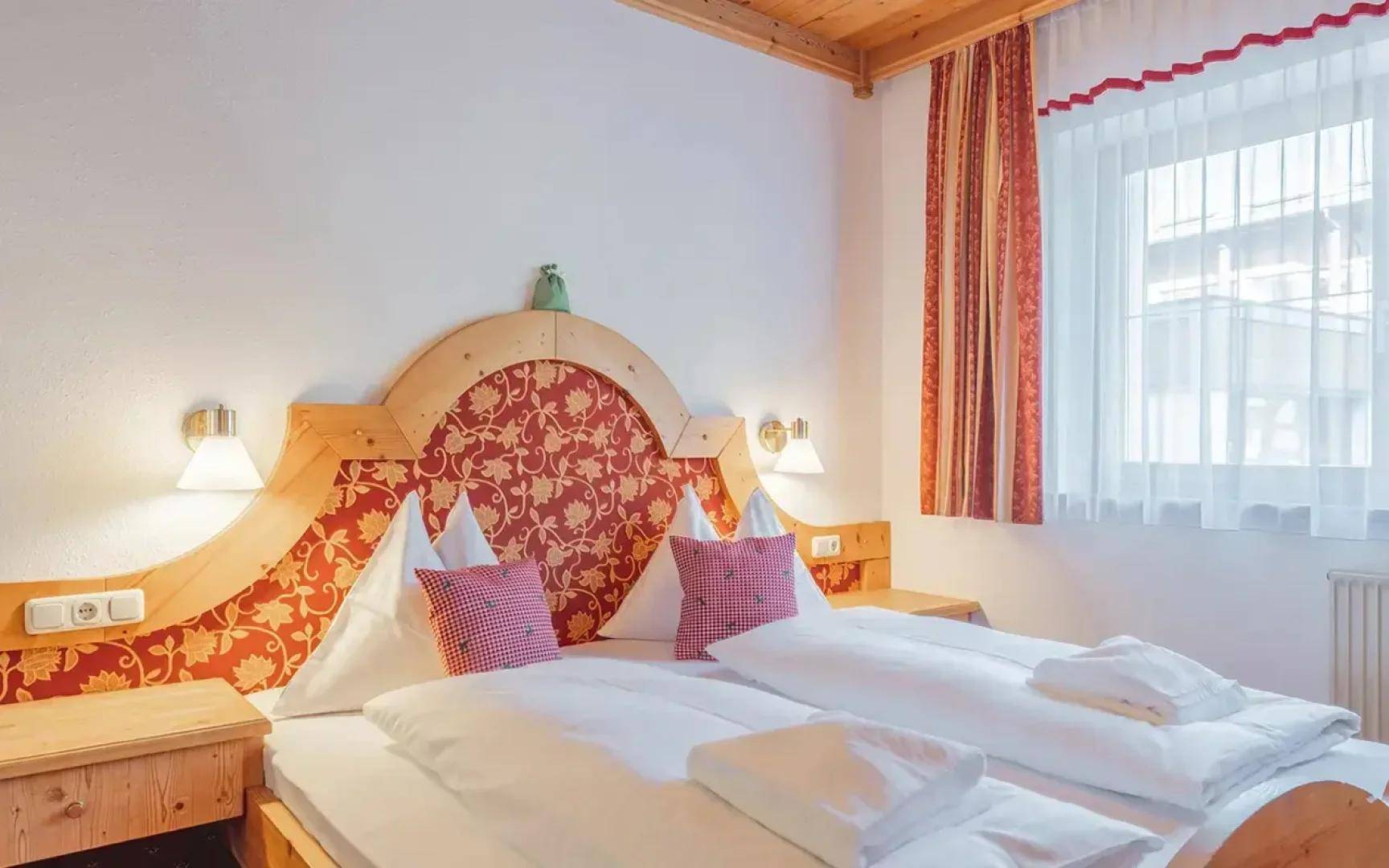 Gemütliches Schlafzimmer mit traditionellem Doppelbett, Holzkopfteil und einem Fenster mit roten Vorhängen.