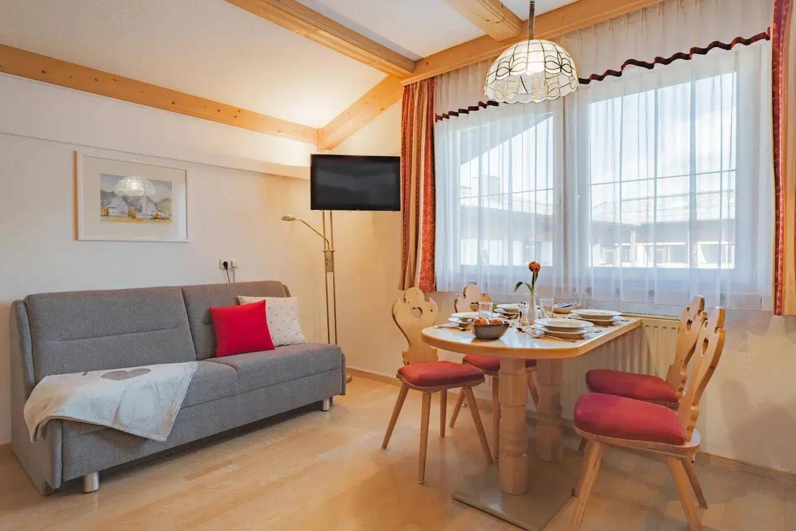Heller Wohn- und Essbereich mit bequemer Couch, Esstisch und großen Fenstern unter Holzbalken.
