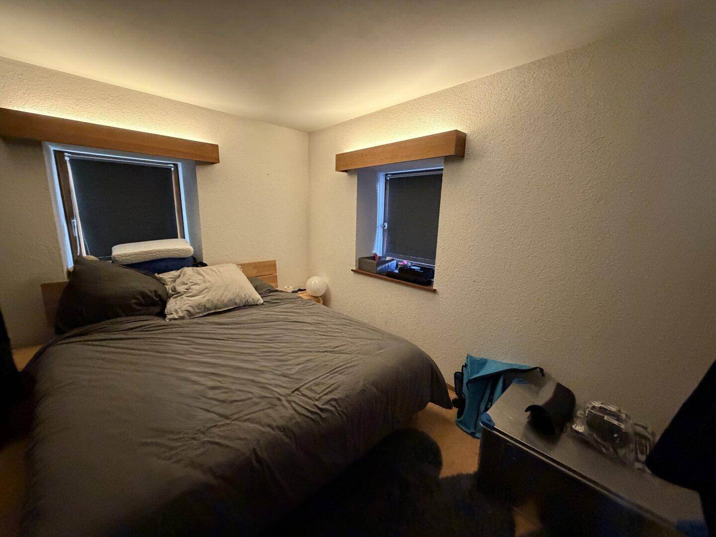 Schlafzimmer mit Doppelbett, zwei Fenstern und indirekter, warmer Beleuchtung.