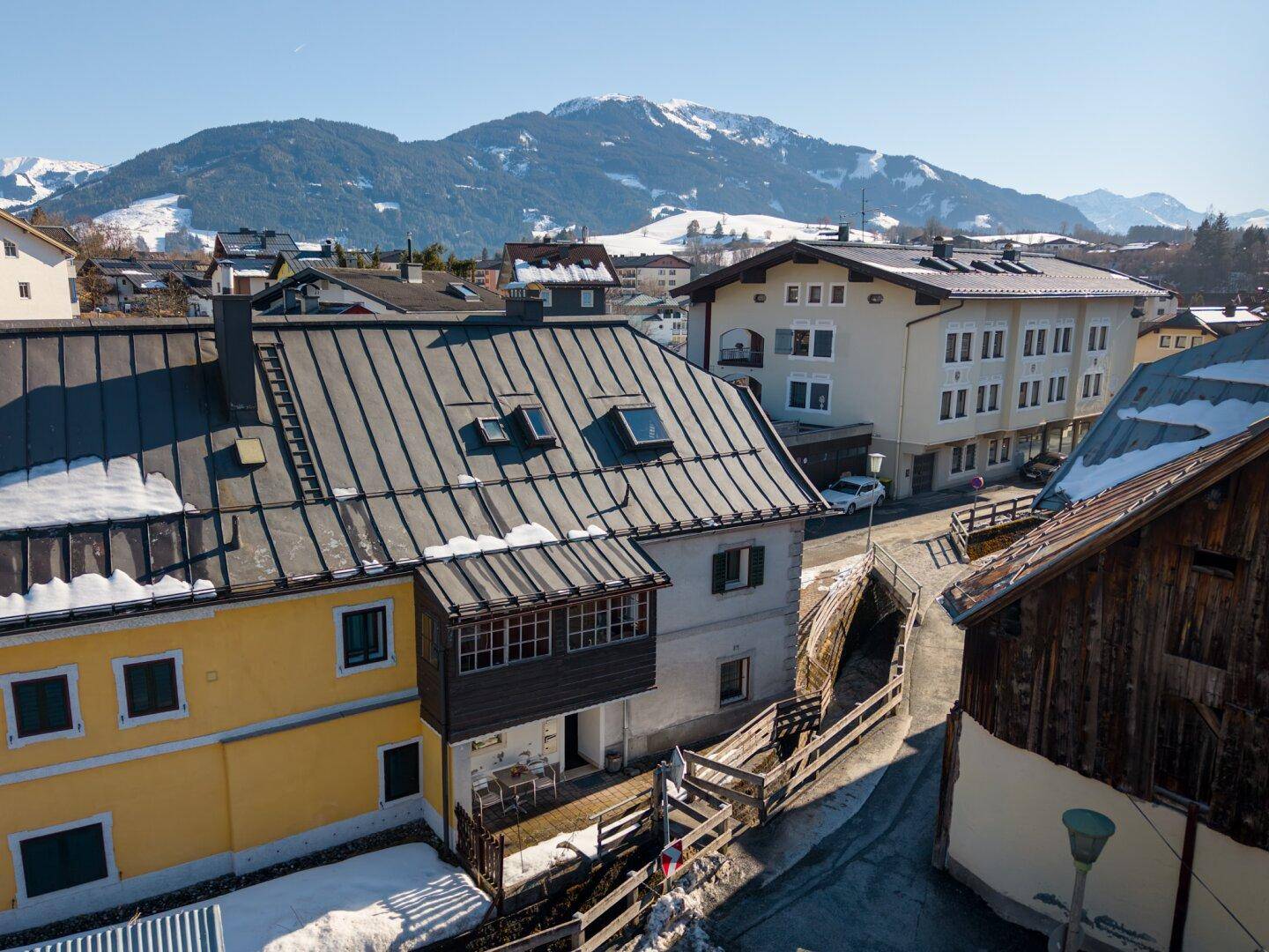 Dachansicht eines Hauses mit umliegenden Gebäuden und schneebedeckten Bergen im Hintergrund.