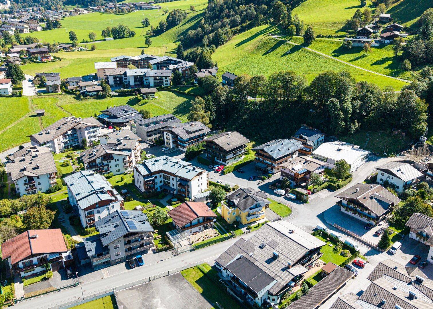 Panoramablick aus der Luft auf ein malerisches Dorf, eingebettet in eine weite grüne Berglandschaft unter blauem Himmel.