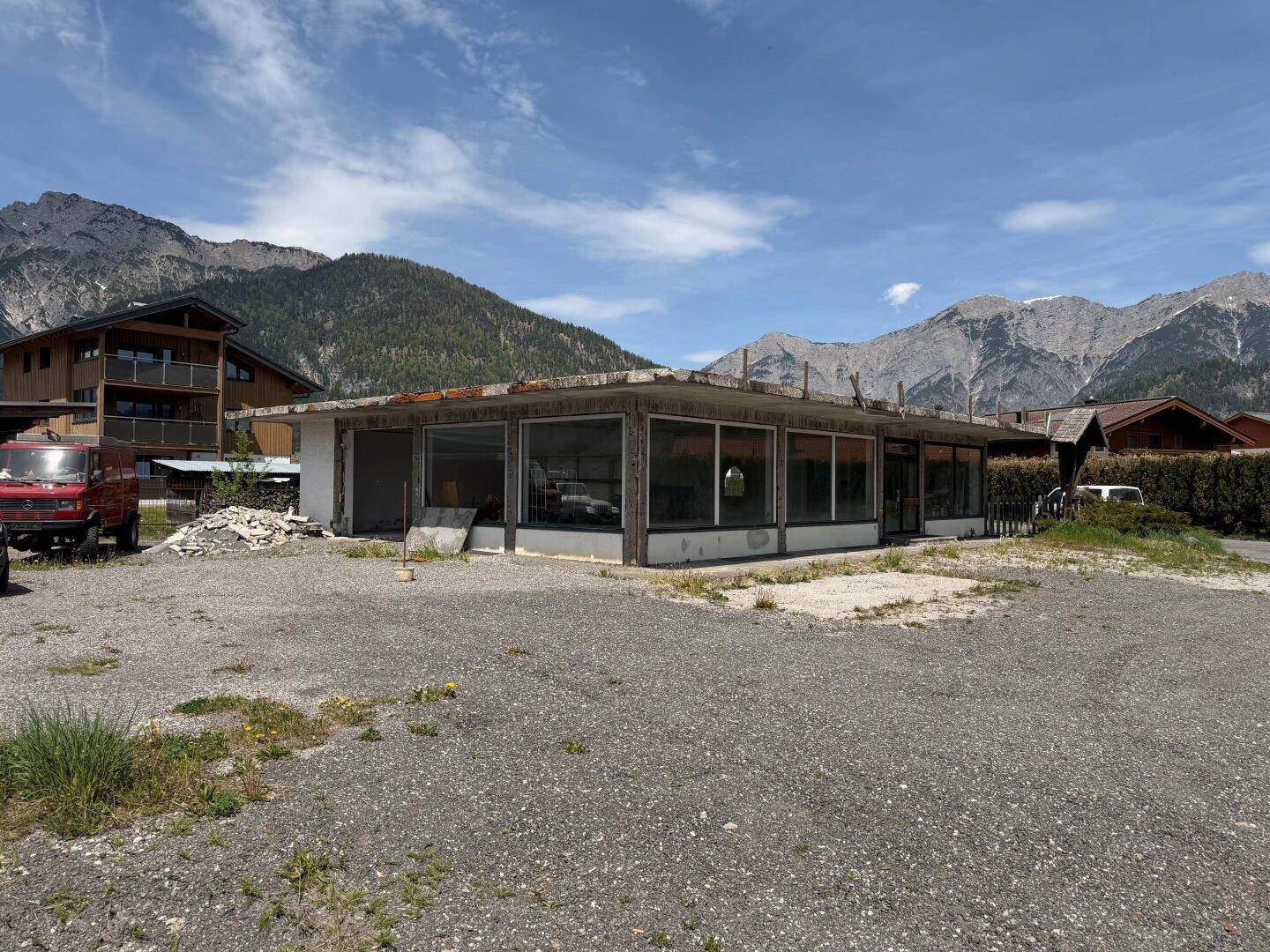 Gebäude im Rohbau mit großen Fensterfronten und Bergblick, umgeben von einer Schotterfläche und Bergen.