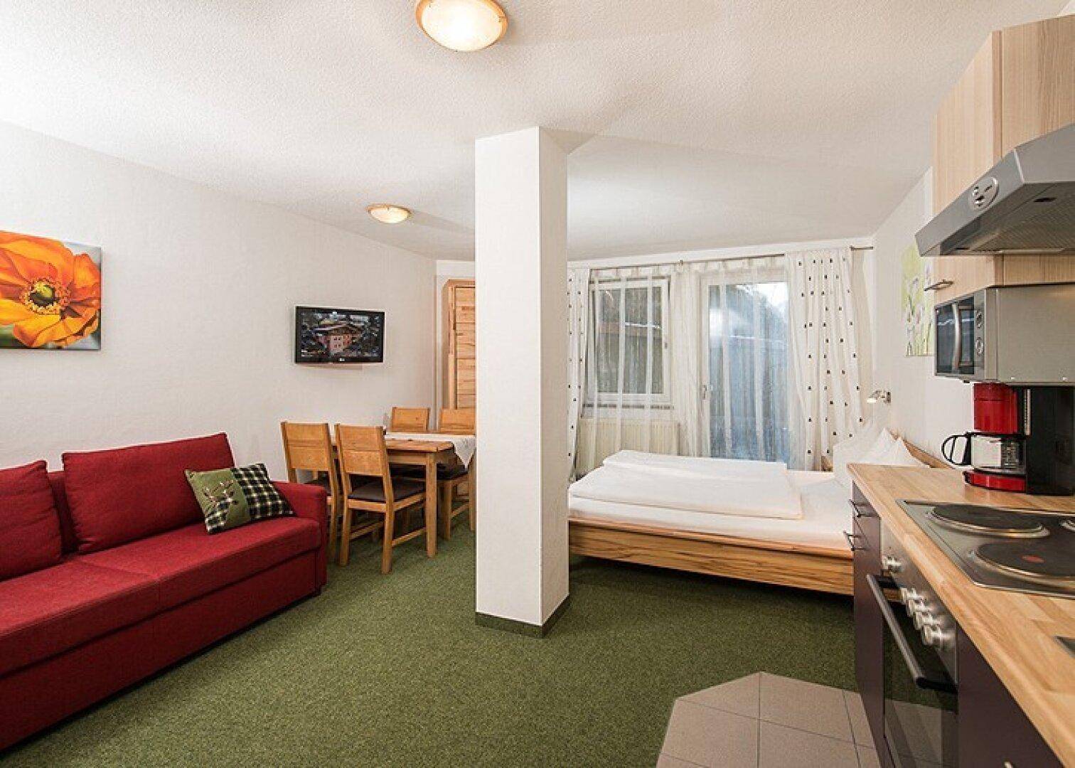 Gemütliches Studio-Apartment mit rotem Sofa, Essbereich, Bett und kleiner Küchenzeile auf grünem Teppichboden.