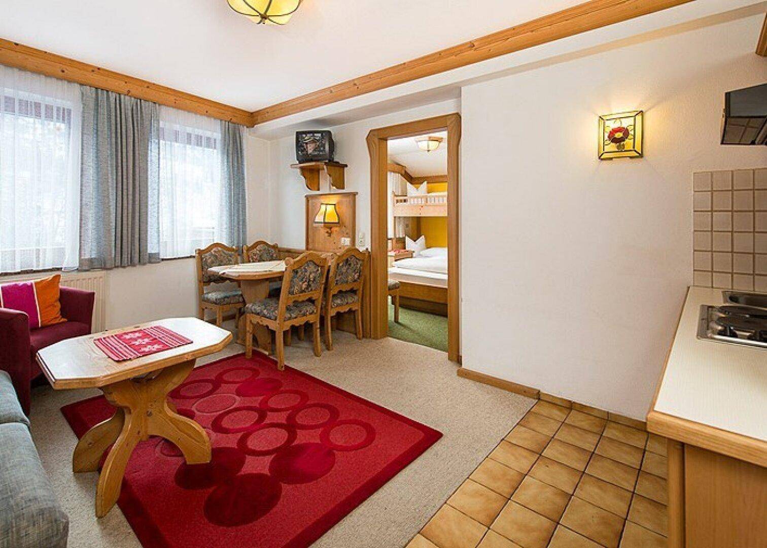 Traditioneller Wohn- und Essbereich mit Holzbalken, rotem Teppich und Zugang zu einem Schlafzimmer mit Etagenbetten.