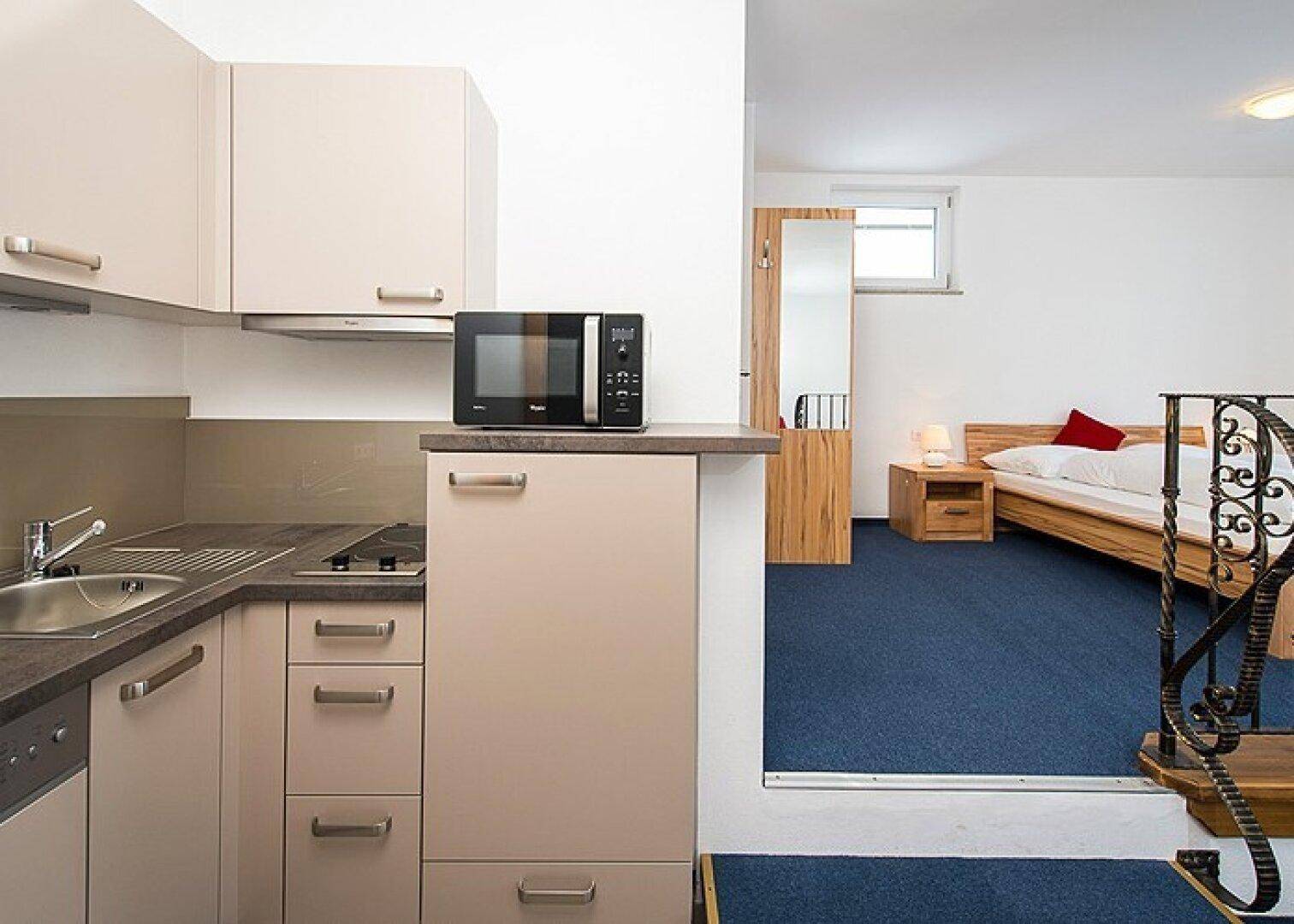Moderne Küchenzeile mit Mikrowelle und angrenzendem Schlafbereich mit Holzbett auf blauem Teppichboden.