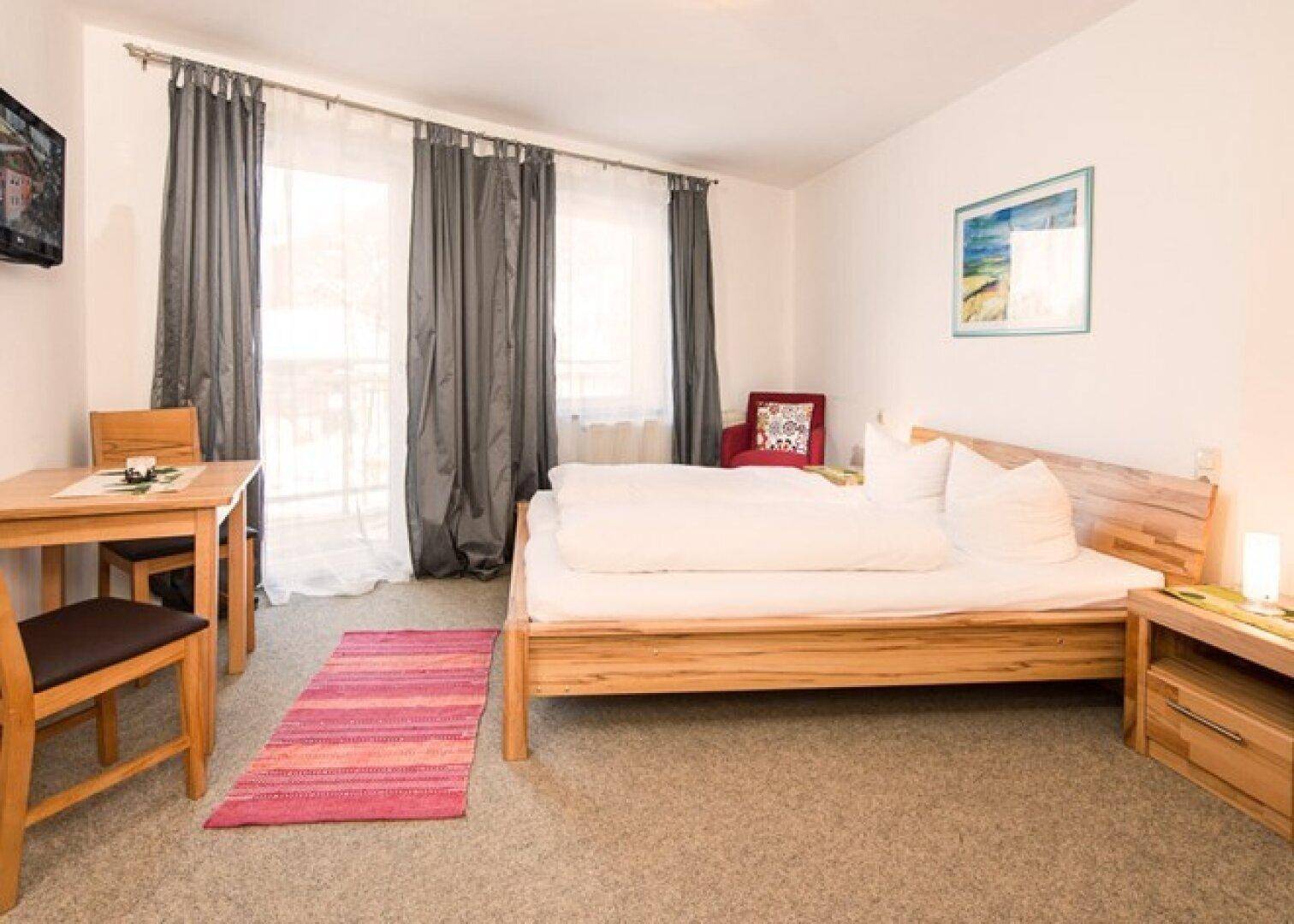 Helles Schlafzimmer mit Holzbett, kleinem Tisch und Stuhl, ausgestattet mit beigem Teppichboden.