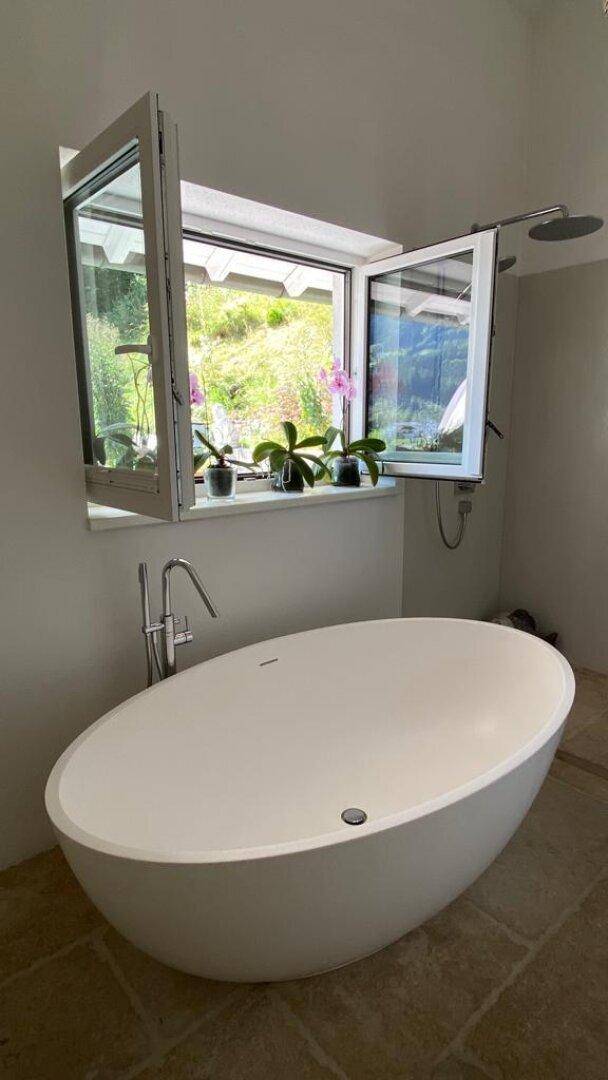 Helles Badezimmer mit freistehender Badewanne, Fenster und Blick ins Grüne.