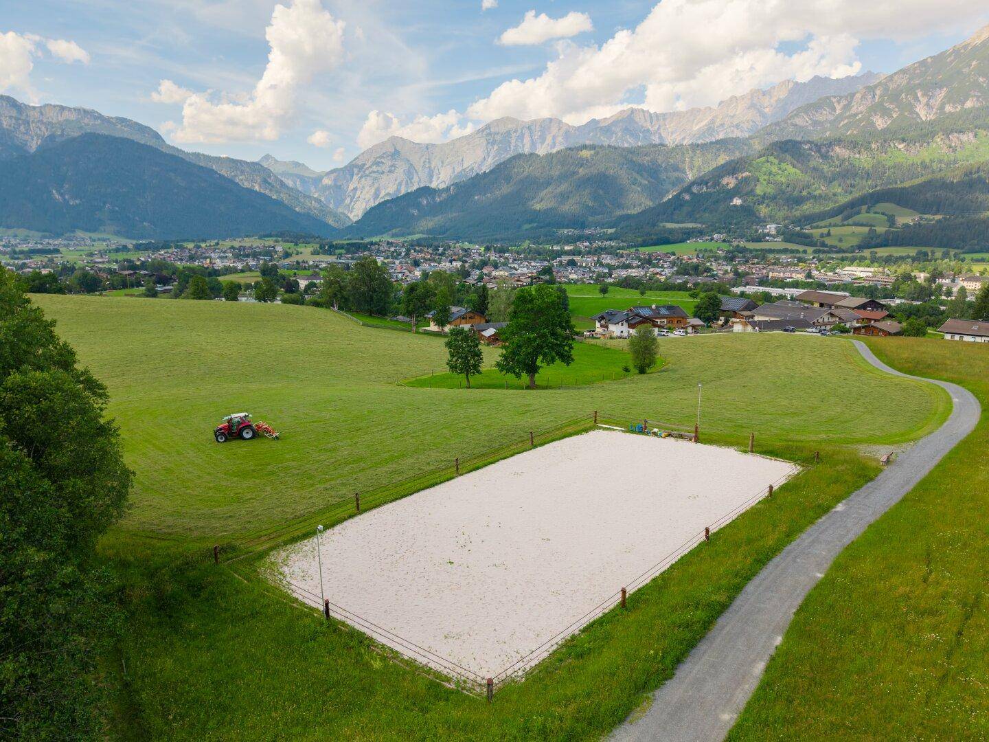 Weitläufiger Reitplatz in ländlicher Umgebung mit Traktor auf der Wiese und Bergblick.