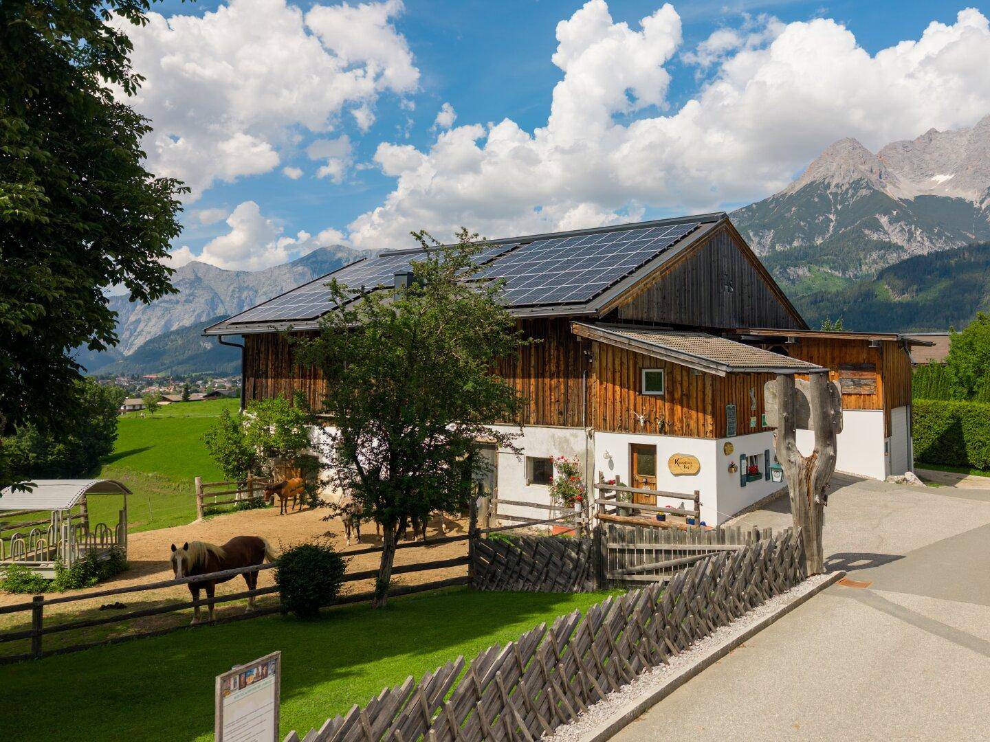 Traditionelles Stallgebäude mit Holzzaun und Pferden, umgeben von grüner Landschaft.