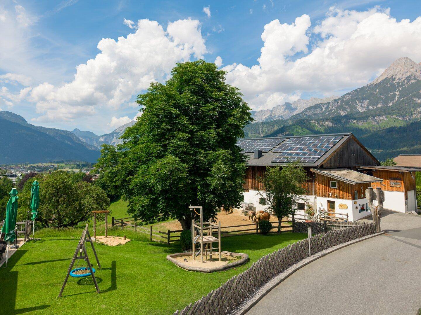 Großer Gartenbereich mit Spielplatz, altem Baum und Blick auf das Stallgebäude und die Berge.