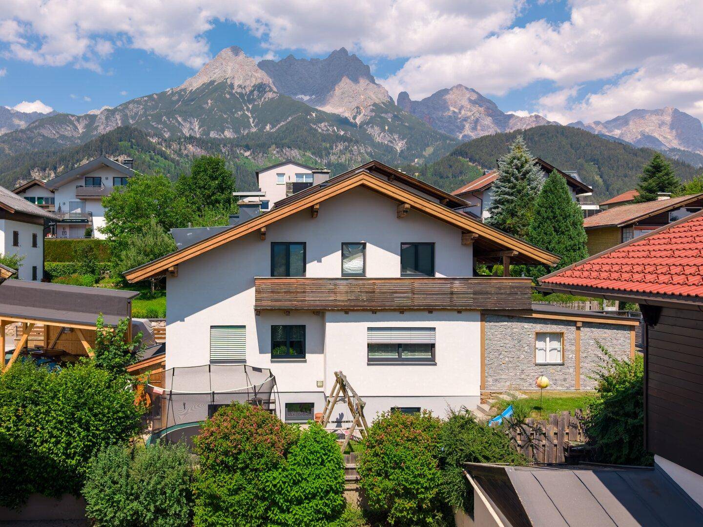 Vorderansicht des Hauses mit Balkon und Spielbereich im Garten, umgeben von beeindruckender Berglandschaft.