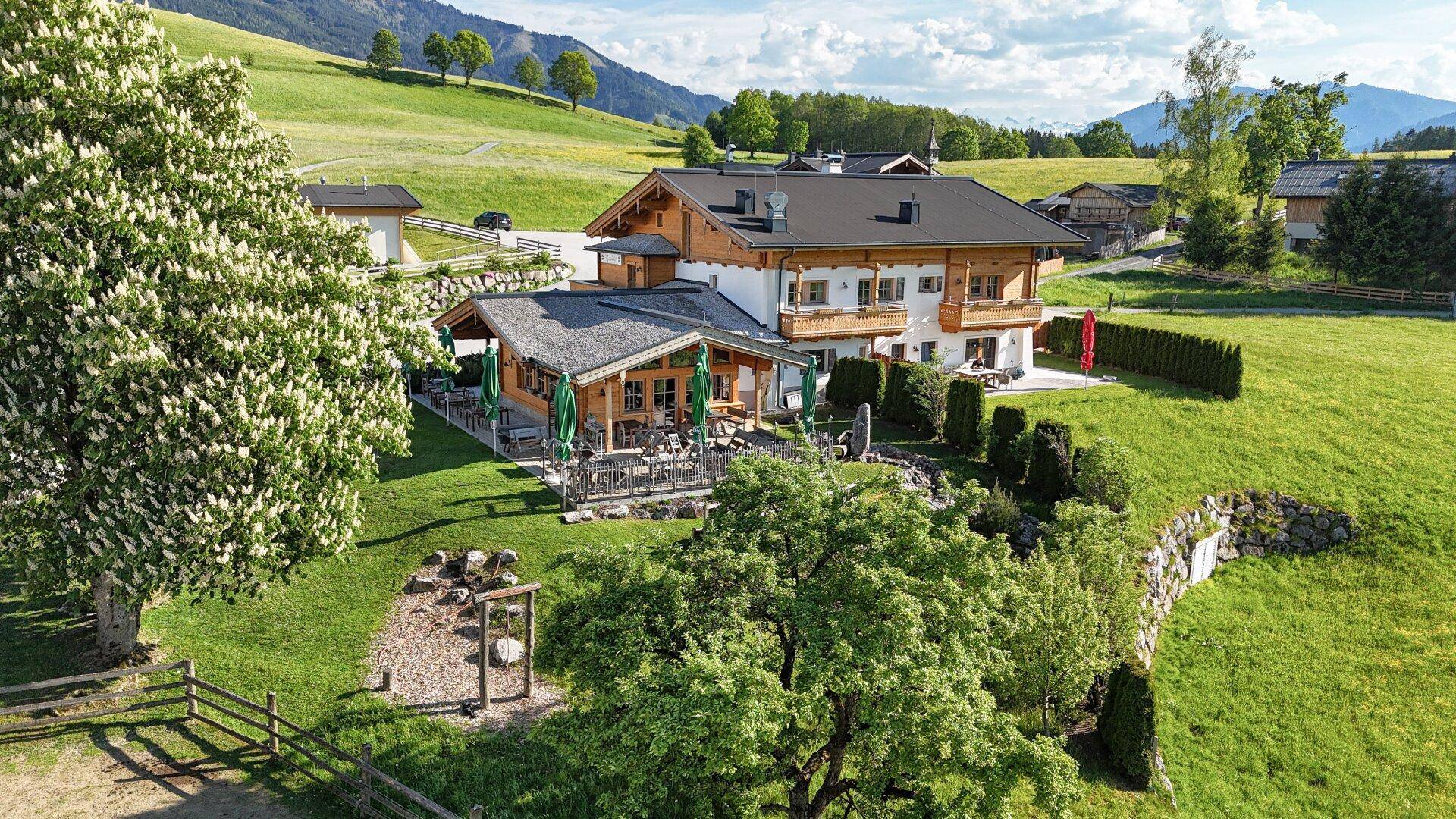 Traditionelles Gebäude mit großer Außenterrasse und Gartenmöbeln, eingebettet in grüne Landschaft.