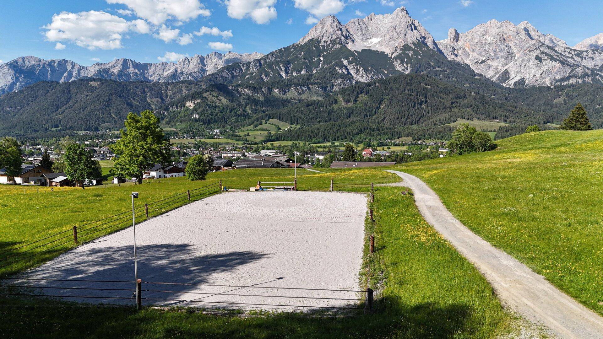 Reitplatz in ländlicher Umgebung mit beeindruckendem Bergpanorama unter blauem Himmel.