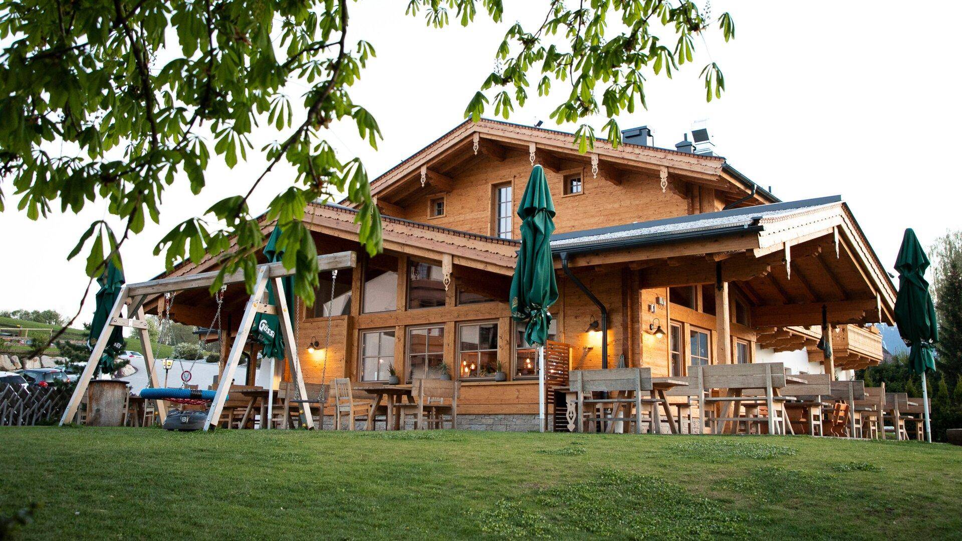 Traditionelles Holzhaus mit Außenterrasse, Schaukel und gepflegtem Rasen unter Bäumen.